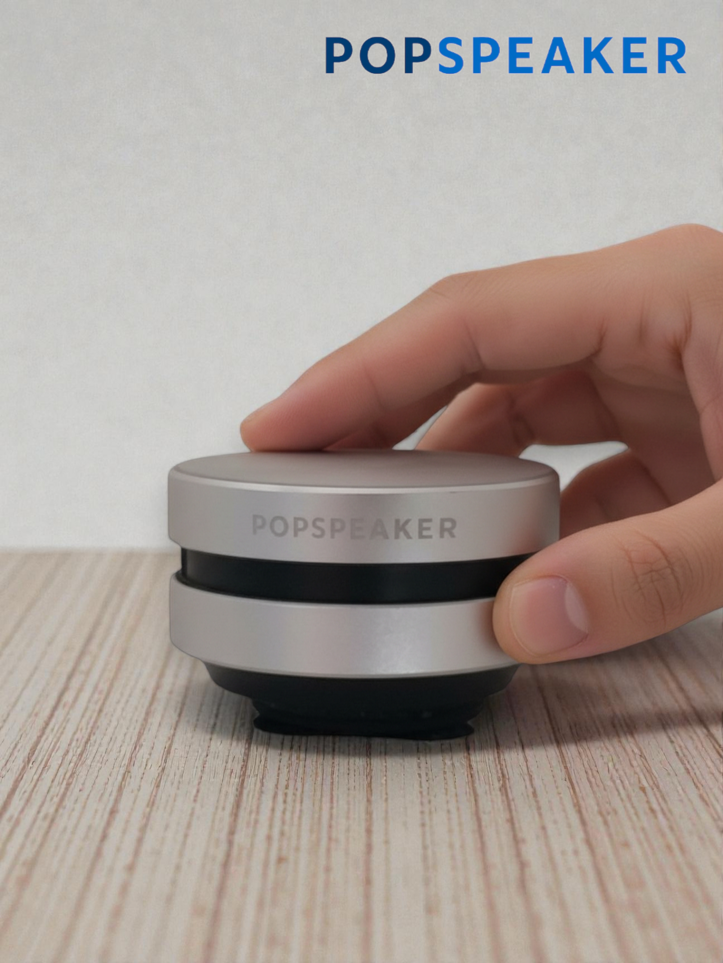 Hand holding a small silver POPSPEAKER device on a wooden surface
